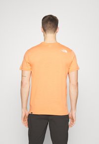 Man with short brown hair wearing an orange The North Face t-shirt and black pants, viewed from behind against a plain background.