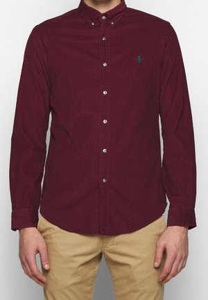 Hombre con una camisa de pana burdeos de manga larga abotonada con un pequeño logo verde bordado en el pecho y pantalones beige.