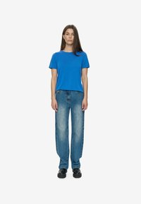Sieviete stāv pretī, ģērbusies zilā T-kreklā, izbalējušās zilās džinsās un melnās masīvās kurpēs uz vienkārša balta fona.