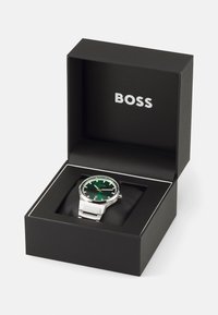 Sudraba rokas pulkstenis ar zaļu ciparnīcu, datuma rādītāju un nerūsējošā tērauda siksniņu melnā kastē ar "BOSS" logo sudrabā.
