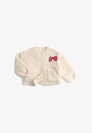 Veste en fausse fourrure crème avec un col arrondi, des boutons-pression, deux poches et un appliqué en forme de nœud rouge sur le côté gauche. Texture douce, coupe décontractée.