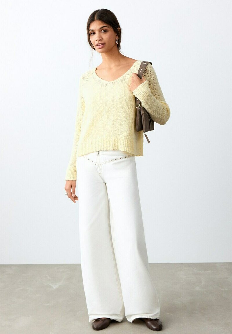 Femme portant un pull en tricot jaune clair, un pantalon large blanc, des chaussures marron et portant un sac à bandoulière marron, debout sur un sol gris.