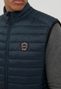 Navy blue padded vest with a high collar, horizontal stitching, and a round logo patch. Worn over a grey long-sleeve shirt.