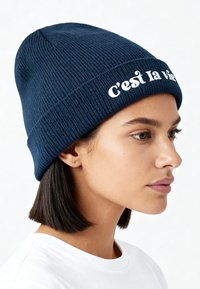 Vrouw met kort donker haar draagt een marineblauwe gebreide beanie met de tekst "C’est la vie" en een wit overhemd, kijkt naar rechts.