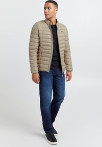 Chaqueta de plumas acolchada beige con cuello alzado, combinada con una camiseta negra y jeans de un azul oscuro. Zapatillas negras completan el conjunto.