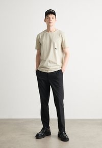 Beige bomullströja med liten logotyp, svarta byxor och svarta skor. En svart keps med "BOSS" i vit text fulländar outfiten.