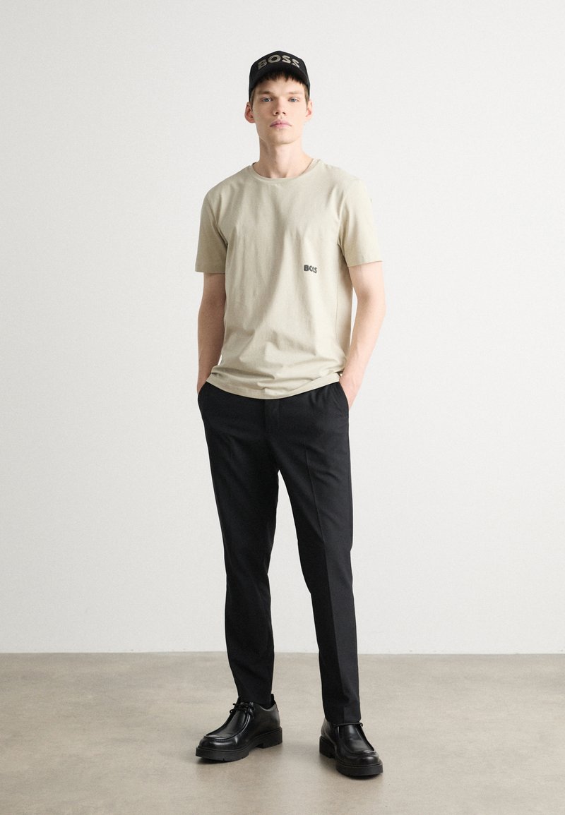 Beige bomullströja med liten logotyp, svarta byxor och svarta skor. En svart keps med "BOSS" i vit text fulländar outfiten.