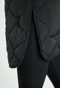 Gilet trapuntato nero in tessuto testurizzato, con uno spacco laterale e bordi arrotondati, indossato sopra pantaloni neri attillati.