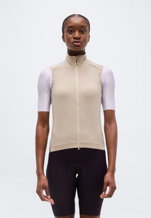 Femeie purtând un vestă bej fără mâneci, cu fermoar, peste un tricou alb cu mânecă scurtă și pantaloni scurți de ciclism negri, privind înainte pe un fundal simplu.