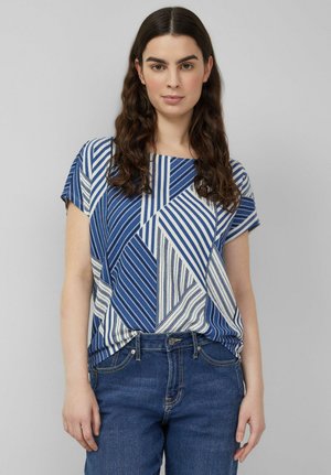 Femme aux longs cheveux ondulés et foncés portant un haut à manches courtes à rayures géométriques bleues et blanches, rentré dans un jean en denim bleu, sur fond neutre.