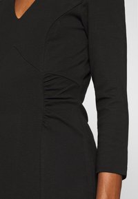 Robe noire avec un décolleté en V, des manches trois-quarts, présentant un ruché sur les côtés et une texture de tissu lisse, mettant en valeur une silhouette ajustée.