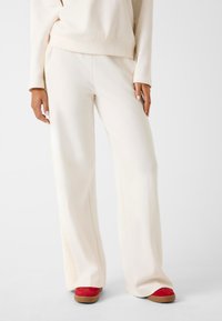 Pantaloni a gamba larga color crema con una texture liscia e accenti a coste sui lati, abbinati a calzature rosse con una suola testurizzata.