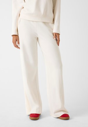 Pantaloni a gamba larga color crema con una texture liscia e accenti a coste sui lati, abbinati a calzature rosse con una suola testurizzata.