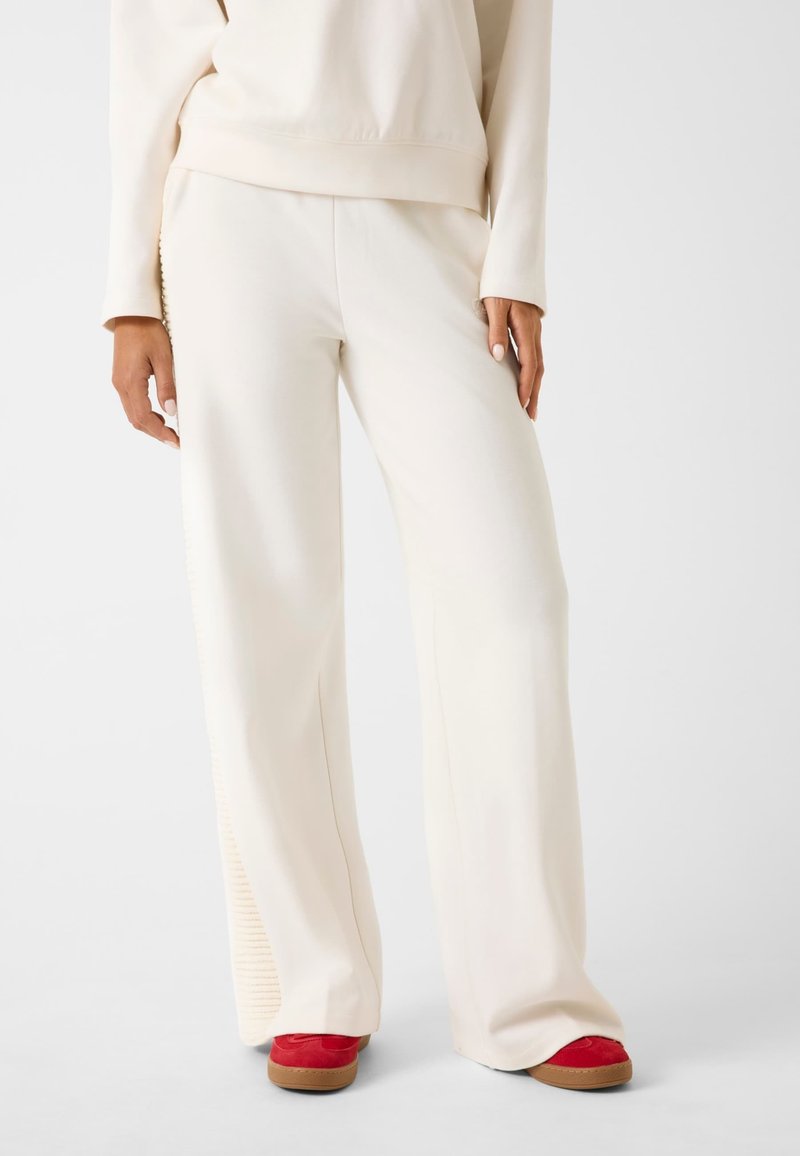 Pantaloni a gamba larga color crema con una texture liscia e accenti a coste sui lati, abbinati a calzature rosse con una suola testurizzata.