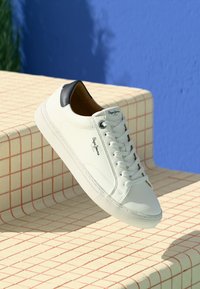 White low-top sneaker with black heel detail and white laces, hovering over beige tiled steps with red grid lines against blue wall.