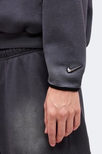 Main et poignet portant un haut à manches longues à carreaux gris foncé avec un logo Nike noir sur le poignet.
