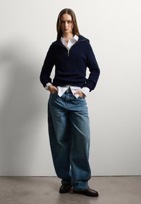 Pull en maille côtelée bleu marine avec un col zippé sur une chemise blanche à boutons, associé à un jean bleu décontracté et des chaussures marron foncé.