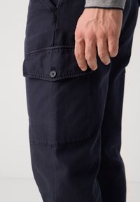 Navy cargo kalhoty vyrobené z odolné bavlny, s kapsou na boku, prošitými detaily a knoflíkovým uzávěrem na kapse pro bezpečné uložení.