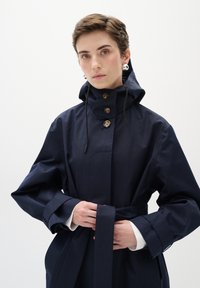 Tumši zila ūdensnecaurlaidīga virsjakas ar kapuci, ar trim brūniem pogām, šņorējamu jostu un platām piedurknēm. Galvējā daļa ir akcentēta ar jostu.