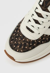 Λευκό αθλητικό παπούτσι με πάνελ από ύφασμα σε καφέ σχέδιο και μεταλλικά τρουκ, δ ιαθέτοντας λευκά κορδόνια και υφασμένη σόλα για καλύτερη πρόσφυση.