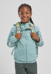 Giacca impermeabile azzurro chiaro con chiusura a zip, polsini elasticizzati e cappuccio, indossata sopra uno zaino verde con spallacci grigi.