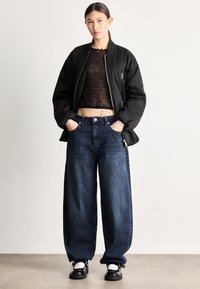 Čierna bomber bunda cez priehľadný čierny krátky top s textom, kombinovaná so širokými tmavomodrými džínsami a čiernymi topánkami s bielymi detailmi.