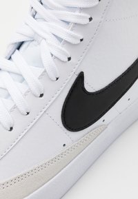 Sneaker in pelle bianca con logo in gomma nera, accentuato da suede grigio testurizzato, lacci bianchi e dettagli perforati vicino alla suola.