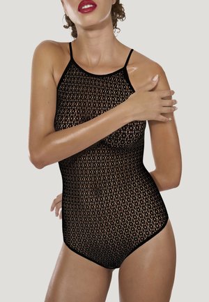 Femme portant un rouge à lèvres rouge et un justaucorps sans manches en résille noire, posant avec un bras croisé sur la poitrine sur un fond uni.