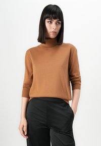 Vrouw met steil zwart haar die een bruine coltrui en zwarte broek draagt, staande met één hand in de zak tegen een witte achtergrond.