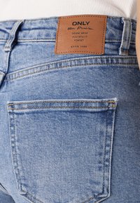 Calças de ganga azul claro com costura laranja, apresentando um patch de couro castanho rotulado "ONLY" na cintura e um design de bolso traseiro.