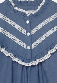 Denimklänning med vita spetsdetaljer och volangkrage. Har fyra knappar och ett zigzag-mönster av spets mellan bröststripes.