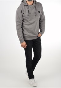 Felpa grigia con cappuccio e coulisse, tasca frontale e piccolo logo sul petto, abbinata a jeans neri e scarpe da ginnastica bianche.