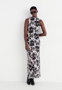 Rochie maxi în imprimeu cu vacă alb-negru, fără mâneci, cu guler înalt. Modelul poartă ochelari de soare negri și botine. Textura țesăturii este netedă.