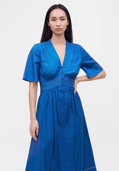 Γυναίκα με μακριά σκουρόχρωμα μαλλιά που φοράει ένα μπλε φόρεμα με κουμπιά και κοντά μανίκια, στέκεται με το ένα χέρι στη μέση της σε λευκό φόντο.