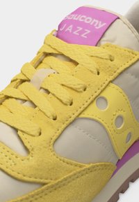 Κίτρινο και μπεζ αθλητικό παπούτσι Saucony Jazz με υφασμάτινα κορδόνια, κίτρινες επενδύσεις από σουέντ και ροζ ετικέτα με λογότυπο στη γλώσσα.
