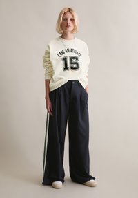 Kremowa bluza z napisem "JESTEM SPORTOWCEM 15" na przodzie. Czarnie spodnie o szerokiej nogawce z białymi paskami bocznymi. Modelka nosi jasne buty.