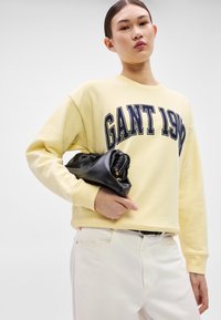 Sárga pulóver "GANT 1949" felirattal, fehér nadrággal és fekete clutch táskával párosítva. A pulóveren kerek nyakkivágás és hosszú ujjak találhatók.