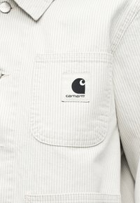 Μπεζ ριγέ σακάκι με εμπρόσθια τσέπη στο στήθος που διαθέτει μαύρο και άσπρο λογότυπο Carhartt.