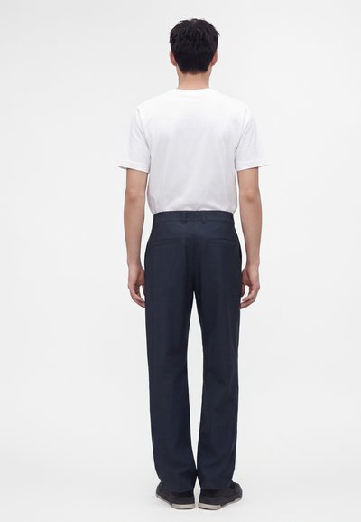 Homme debout de dos portant un t-shirt blanc uni, un pantalon droit bleu foncé et des chaussures noires sur fond blanc.