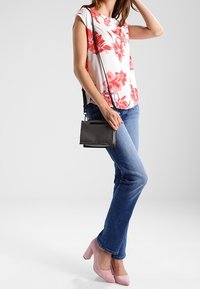 Blusa floral de manga corta con flores rojas sobre fondo blanco, combinada con vaqueros azules, zapatos de tacón altos rosas y un pequeño bolso bandolera negro.