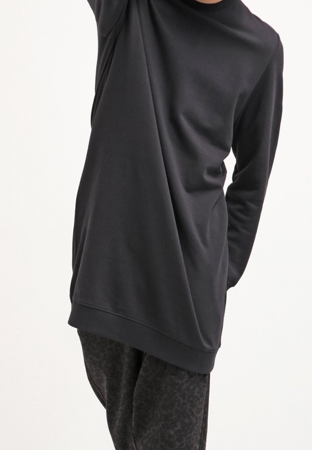Personne portant une chemise noire ample à manches longues et un pantalon foncé à motifs, levant un bras au-dessus de la tête, sur un fond uni clair.