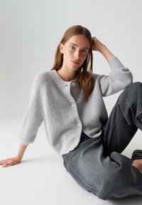 Jeune femme aux longs cheveux bruns portant un cardigan gris clair à boutons et un jean sombre ample, assise contre un fond clair uniforme.