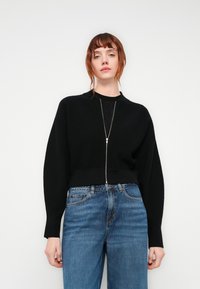 Svart cropped genser med ribbestruktur, med glidelås foran og videmontringer, kombinert med høyhalsede blå jeans.