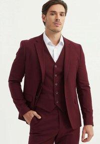 Costume trois pièces bordeaux avec un blazer ajusté, un gilet assorti doté de boutons et un pantalon sur mesure. Chemise blanche en dessous.