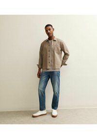 Giacca camicia beige con bottoni, t-shirt grigia sotto, jeans blu strappati e sneakers bianche con suole marroni. In piedi su uno sfondo neutro.