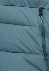 Vadderad jacka i ljusblått tyg med en slät textur. Har en synlig dragkedja längs sidan och horisontella sömmar.