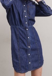 Jeansová šaty v tmavě modré barvě s knoflíkovým zapínáním vpředu, límcem a elastickým pasem. Obsahují náprsní kapsu a dlouhé rukávy s knoflíkovými manžetami.