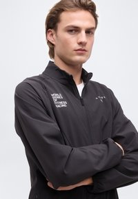 Νεαρός άνδρας με σταυρωμένα χέρια που φοράει μαύρο μπουφάν με φερμουάρ, με τα λογότυπα «World Series of Fitness Racing» και «Hyrox» στο μπροστινό μέρος