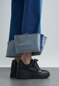 Černé kotníkové tenisky na klínku s kombinací hladkých a strukturovaných materiálů. Kombinovány s ohrnutými džínovými kalhotami, které ukazují kontrast světle a tmavě modré.