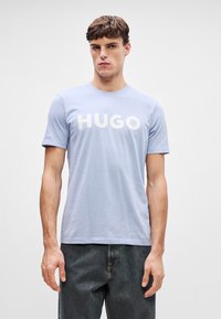 Ανοιχτό μπλε βαμβακερό t-shirt με κοντά μανίκια, που έχει τη λέξη "HUGO" σε έντονους λευκούς χαρακτήρες στη διάρκεια του στήθους. Απλή σχεδίαση με στρογγυλή λαιμόκοψη.